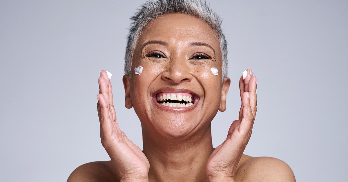 Image shows happy mature woman with little dabs of face cream on each cheek.