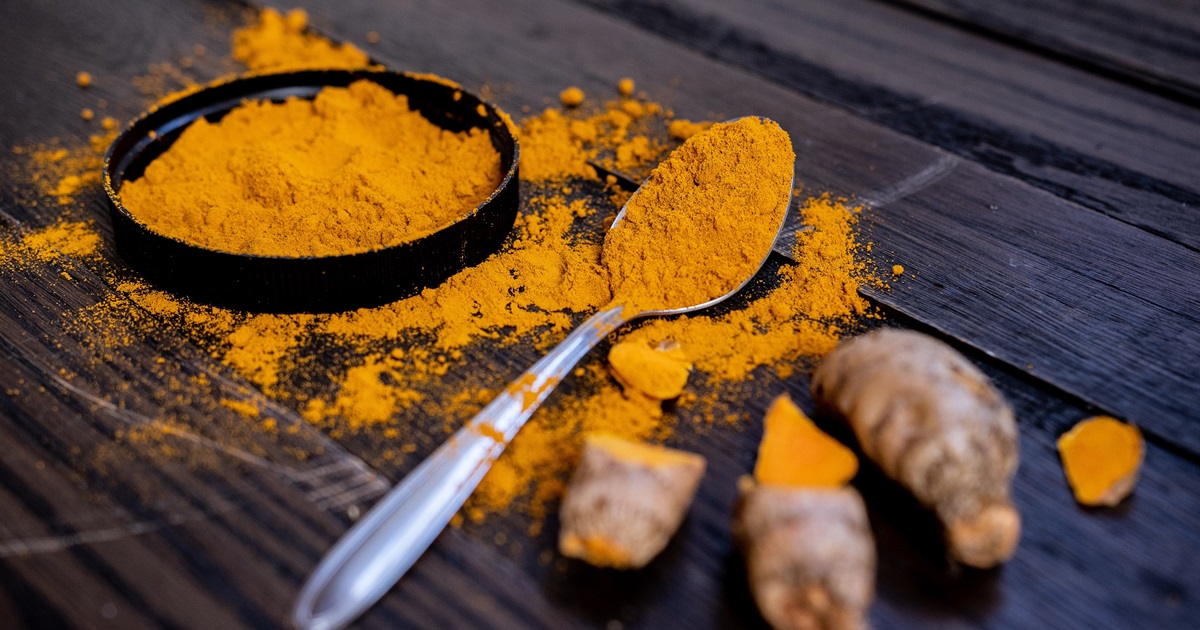 Image shows bowl of turmeric powder on a black table top with turmeric roots around